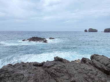 Okyanus dalgalarının bulutlu bir gökyüzünün altındaki kayalara çarptığı manzaralı kayalık kıyı şeridi. Seyahat, doğa ve çevre temaları için uygun doğal deniz manzarası.