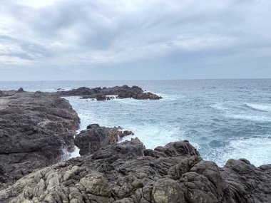 Okyanus dalgalarının bulutlu bir gökyüzünün altındaki kayalara çarptığı manzaralı kayalık kıyı şeridi. Seyahat, doğa ve çevre temaları için uygun doğal deniz manzarası.