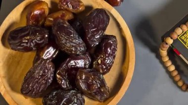 Kurumuş hurmaların, dua boncuklarının yanına koyulmuş ahşap bir kâse ve karanlık bir yüzeye Kur 'an' ın sinematik görüntüleri. Sahne Ramazan geleneklerini, oruç tutmayı ve İslam dini uygulamalarını sembolize ediyor. Ses olmadan düzgün kamera hareketi.