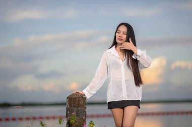 Genç bir kadın gün batımında suyun kenarında zarif bir şekilde poz verir, beyaz bir gömlek giyer, doğal ve huzurlu bir ortamda güzellik ve dinginlik yayar..