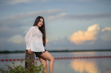 Beyaz gömlekli genç bir kadın, gün batımında sakin sularda zarif pozlar veriyor, sakin bir ortamda doğanın güzelliğini ve sıradan modasını sergiliyor..