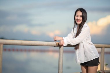 Personel üniformalı genç bir kadın, deniz kenarında güzel bir anın tadını çıkarıyor. Bulutların ve arka planda sakin bir gölün olduğu sakin bir doğa sahnesi sergiliyor..