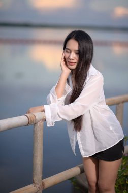 Saf gömlekli genç bir kadın resim gibi bir gölün kenarında poz veriyor, sükunet ve doğanın güzelliğini sergiliyor, turizm ve yaşam tarzı temaları için mükemmel..