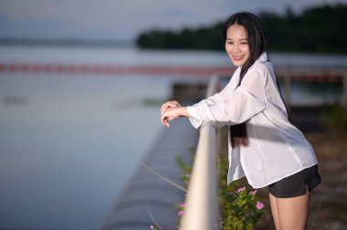 Suyun kenarındaki doğal güzelliğin tadını çıkaran genç bir kadın parmaklıklara yaslanırken bir mutluluk ve rahatlama anı yakalıyor..