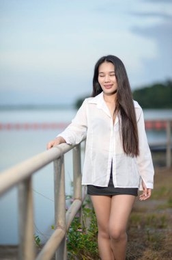 Beyaz gömlekli genç bir kadın, deniz kenarında sakin bir anın tadını çıkarıyor, turizm ve doğa titreşimlerini somutlaştırıyor. Manzara sükuneti ve güzelliği vurguluyor..