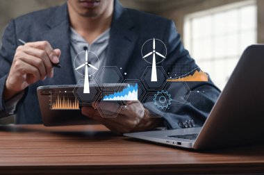 Bir profesyonel, tablet ve dizüstü bilgisayar kullanarak rüzgar türbinleriyle ilgili yenilenebilir enerji verilerini değerlendirir. Grafik temiz enerji çözümlerini destekleyen analitikleri temsil ediyor.