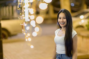 Neşeli genç bir kadın sıcak bir şekilde gülümser. Parlak dekoratif ampullerin altında durur. Açık havada, davetkar bir arka planda..