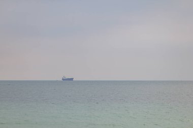 Bulutlu bir gökyüzüne karşı uzak bir kargo gemisini tasvir eden sakin bir deniz manzarası. Sakin su mavi ve yeşilin tonlarını yansıtarak barışçıl bir atmosfer yaratır..