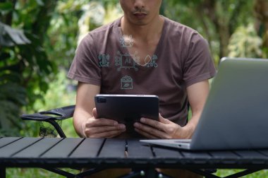 Bir adam yemyeşil bir bahçede oturuyor, tarım alanındaki yapay zeka uygulamalarını keşfetmek için tablet kullanıyor. Dijital simgeler tarım teknolojisindeki çeşitli yenilikleri temsil ediyor.