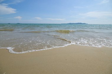 Bu görüntü, Tayland 'ın Rayong Eyaleti' ndeki sakin bir plaj sahnesini yakalıyor. Açık mavi gökyüzünün altında kumlu kıyıya doğru hafif dalgalar süzülüyor..