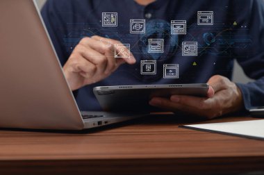 Modern bir çalışma alanında bir kişi dizüstü bilgisayar kullanırken bir tabletle meşgul olur. Resim, bağlantı ve teknolojiyi gösteren dijital elementlerle etkileşimi gösteriyor..