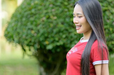 Genç bir kadın dışarıda dikiliyor, günlük kırmızı bir gömlek giyiyor, neşeli bir gülümseme sergiliyor. Yeşil yapraklarla çevrili, mutluluk ve sükuneti temsil ediyor..