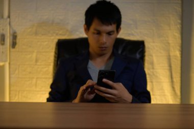 Siyah takım elbiseli genç bir adam akıllı telefonuna dalmış bir şekilde masada oturuyor. Sıcak iç aydınlatma, modern teknolojiyi ve profesyonel davranışı vurgulayan davetkar bir atmosfer yaratıyor..