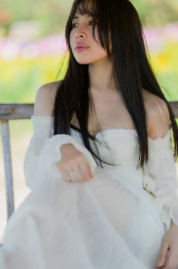 Güzel bir kadın rengarenk çiçeklerle çevrili bir bahar tarlasında zarifçe oturur. Yumuşak güneş ışığı, onun huzurlu ifadesini vurgular, doğadaki bir sükunet anını yakalar..