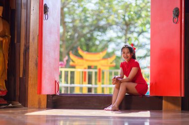 Kırmızı giyinmiş neşeli bir kız güneşli bir kapı eşiğinde oturuyor ve canlı bir tapınakta yeni yılı kutluyor. Neşeli ifadesi festival sezonunun ruhunu yansıtıyor..