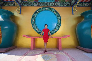 Çarpıcı kırmızı elbiseli genç bir kadın canlı, renkli bir arka plana karşı güven içinde poz veriyor, geleneksel Çin dekoru sergiliyor..