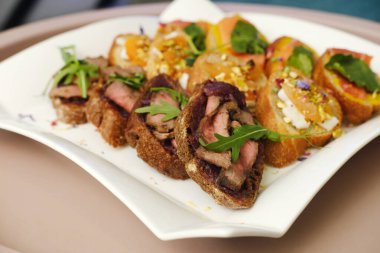 Izgara et ve taze otlarla süslenmiş çeşitli gurme kanepeler sofistike bir mutfak gösterisi için beyaz bir tabakta zarif bir şekilde sunuluyor.