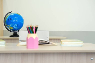 Sınıf ortamında bir dünya, pembe bir tutacakta renkli kalemler ve ahşap bir masanın üzerinde açık kitaplar gösterilerek eğitici bir atmosfer yaratılıyor.