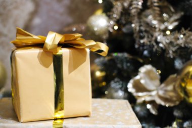 Zarif altın bir hediye kutusu. Masasında parlak bir kurdele duruyor. Etrafı şenlikli süslemelerle ve güzel süslenmiş Noel ağacıyla çevrili.