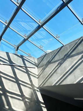 Güneş ışığının cam tavandan süzülen, gri duvarlarda çarpıcı geometrik gölgeler yaratan, modern mimari tasarım ve ambiyansı vurgulayan...