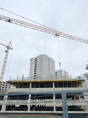 İnşaat alanı, bulutlu bir gökyüzünün altında inşaat iskelesi olan vinçleri ve bitmemiş konut binalarını gösteriyor, kentsel gelişimi ve altyapı ilerlemesini vurguluyor