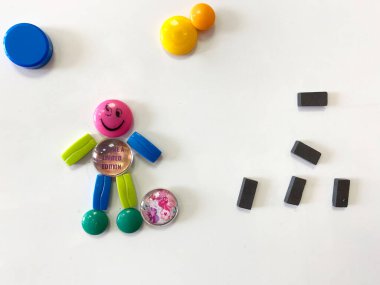 Beyaz bir yüzeyde, plastik parçalardan ve küçük dikdörtgen mıknatıslardan oluşan gülümseyen bir karakter de dahil olmak üzere renkli manyetik şekiller ve şekiller düzenlenmiştir.