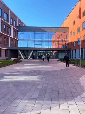 Camdan önü ve canlı duvarları olan modern eğitim tesisi, öğrenciler güneşli bir açık hava ortamında asfalt bir yolda yürüyor.