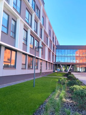 Modern ticari bina büyük cam pencereler ve yeşil peyzajlar içeriyor, parlak mavi gökyüzü altında doğal ışık altında çağdaş mimariyi vurguluyor.