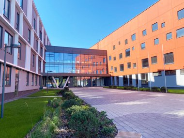 Modern okul binasında geniş cam bir giriş ve peyzaj alanlarıyla çevrili renkli bir dış görünüş ve berrak mavi bir gökyüzünün altında asfalt bir avlu yer alıyor.