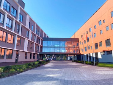 Çok katlı binada geniş cam bir giriş ve peyzaj alanı ve berrak mavi gökyüzü ile çevrili renkli bir cephe yer alıyor ve modern mimariyi gözler önüne seriyor.