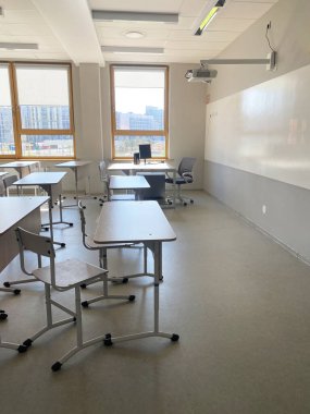 Boş minimalist sınıf, düzgün sıralanmış masa ve sandalyeler, doğal ışık sağlayan büyük pencereler, tavana monte edilmiş projektör ve duvarda beyaz tahta içerir.