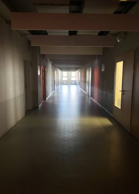 Sonunda parlak ışık olan uzun bir koridor, beyaz duvarlar, ahşap kapılar, ve hareketler için geniş ve davetkar bir ortam sağlayan düz bir zemin.