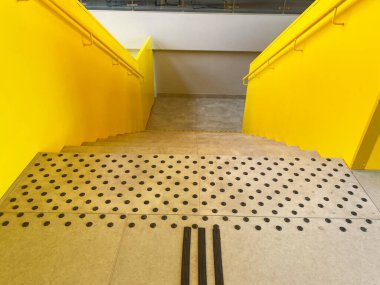 Parlak sarı merdivenler, güvenlik için dokunsal asfalt döşeme ile iniyor, korkuluklar ve çağdaş bir iç dekorun parlak beton zeminiyle tamamlanıyor.