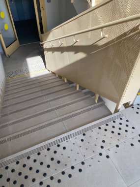 Erişilebilirlik için tasarlanmış merdivenler, dokunsal işaretler ve tırabzan ile, kapıları olan bir koridora açılıyor, doğal ışıkla aydınlatılıyor.