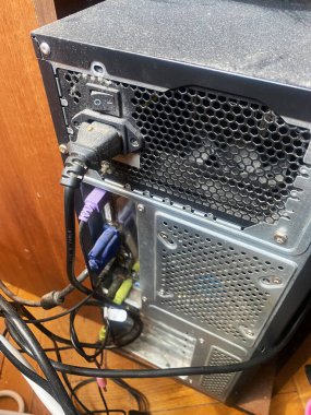 Güç kaynağı bağlantılarını, birden fazla kabloyu ve yüzeyinde toz ve ahşap zemin olan bir havalandırma ızgarasını gösteren bir bilgisayar kulesinin arka görüntüsü.