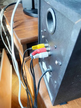 Siyah hoparlör, ahşap bir masa üzerinde bağlı kablolar ile RCA giriş portlarını özelleştirir, diğer elektronik aygıtlar arka planda kısmen görünür