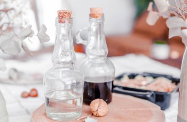 Mantar tıpalı cam şişeler, biri berrak sıvı ve diğeri koyu sıvı, ahşap tahtada dekoratif elementlerle sıralanmış, rahat bir atmosfer yaratıyor.