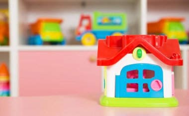 Kırmızı çatılı ve mavi kapılı parlak renkli bir oyuncak evi, bir anaokulu sınıfındaki pembe bir masanın üzerinde, arka planda çeşitli oyuncaklar var.