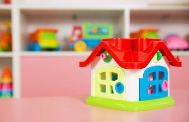 Pembe bir masanın üzerinde kırmızı çatılı, mavi kapılı parlak renkli bir oyuncak ev. Anaokulu sınıfının arka planında eğitici oyuncaklar var.