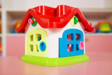 Kırmızı çatısı ve çeşitli kapıları olan parlak renkli plastik oyuncak evi, arka planda oyuncaklar olan pembe bir dolaba yerleştirilmiş.