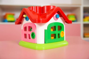Kırmızı çatısı ve yeşil tabanı olan parlak renkli plastik oyuncak evi, çocuk oyun alanındaki pembe bir dolabın önünde sergilenen kapı ve pencereler.