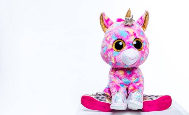 Canlı, yumuşak oyuncak tek boynuzlu at, renkli kürklü ve büyük gözlü, beyaz arka planda konumlanmış, çocukların eğlenmesi için neşeli ve kaprisli tasarımını vurguluyor.