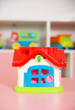 Kırmızı çatısı ve mavi kapısı olan parlak renkli bir oyuncak evi. Arka planda bulanık çocuk oyuncakları olan pembe bir masaya yerleştirilmiş.