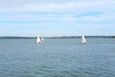 Minsk, Belarus - 30 Nisan 2025: Yelkenliler, uzak tepeler ve açık mavi gökyüzü ile çevrili sakin göl sularında gezinerek, huzurlu bir yaz ortamı yaratıyorlar.