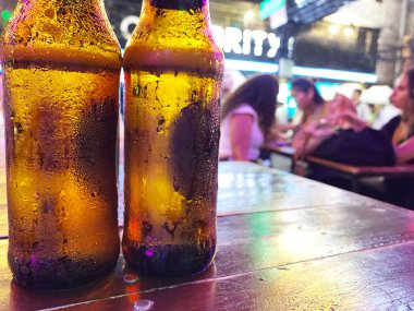 İki soğuk kehribar bira şişesi, ahşap bir masanın üzerinde damlacıklar ve bulanık bir arka plan, insanların canlı bir bar atmosferinde içkiden zevk aldığını gösteriyor.