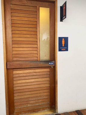 Yatay kirişleri ve cam paneli olan ahşap kapı, halka açık bir alanda, itme kolu olan erkekler tuvaleti tabelasıyla işaretlenmiş.