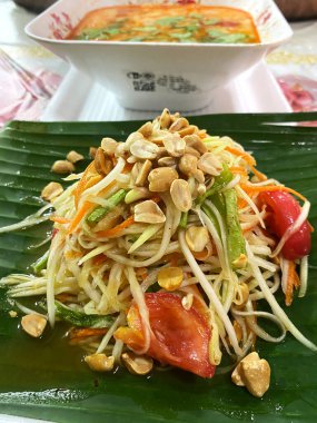 Pad Thai salatası içinde doğranmış sebzeler, fıstıklar ve muz yaprağının üzerinde sunulan otlar ve yanında arka planda bir kase çorba var.