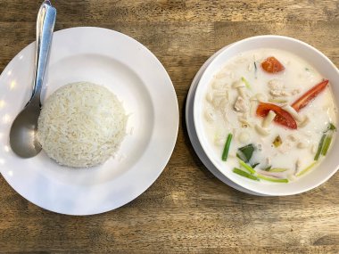 Hindistan cevizi Tom çorbası yuvarlak bir tabakta beyaz pirinç servisinin yanında sunuluyor. Tahta bir yüzey üzerinde pirincin yanında bir kaşık duruyor.