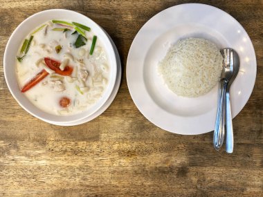 Hindistancevizi Tom çorbası beyaz bir kâse içinde sunuluyor. Yanında buğulanmış pirinç servis ediliyor. Tahta bir yüzey üzerinde çatal ve kaşıkla.