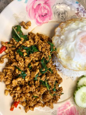 Bir tabakta fesleğen ve baharatlı kızarmış tavuk, yanında da kızarmış yumurta ve salatalık dilimleri, canlı renkler ve dokular.